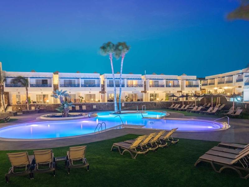 Beleuchteter Pool mit Liegestühlen und zweistöckigem Hotel im Hintergrund bei Dämmerung.