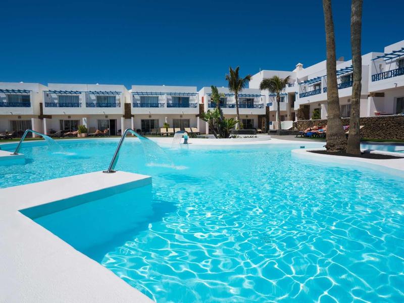 Großer blauer Pool mit weißen Gebäuden und Palmen unter hellem Himmel.