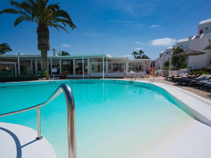 Klares türkisfarbenes Schwimmbecken mit Edelstahlgeländer, Palmen und weißen Gebäuden im Hintergrund.