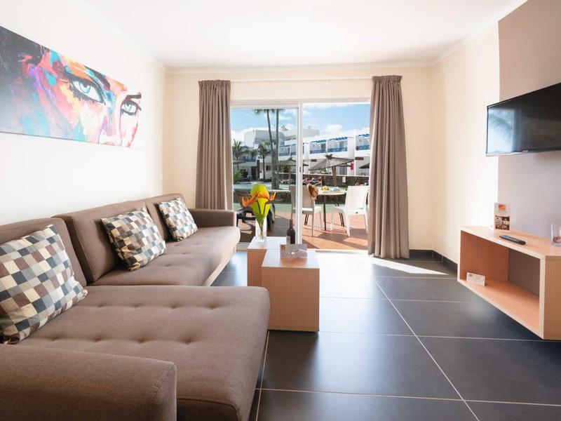 Helles Wohnzimmer mit grauem Sofa, bunten Kissen, Flachbildschirm und Zugang zur Terrasse.