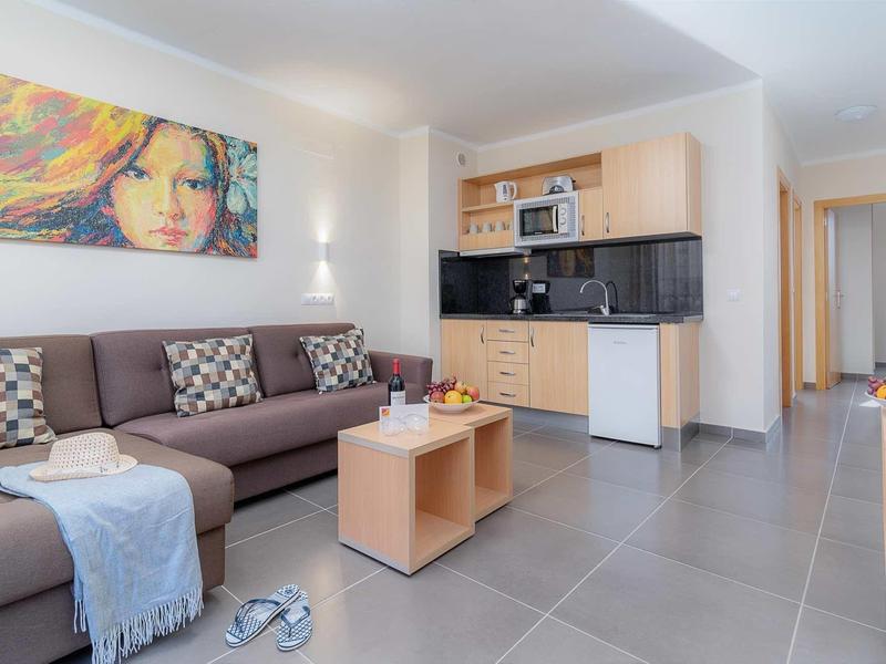 Helles Wohnzimmer mit brachem Sofa, Kissen, modernem Küchenbereich und buntem Wandbild.