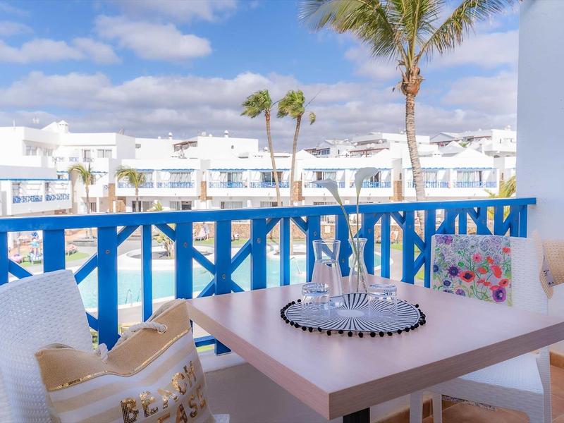 Balkon mit blauem Geländer, weißen Möbeln, Kissen und Palmen bei sonnigem Himmel.