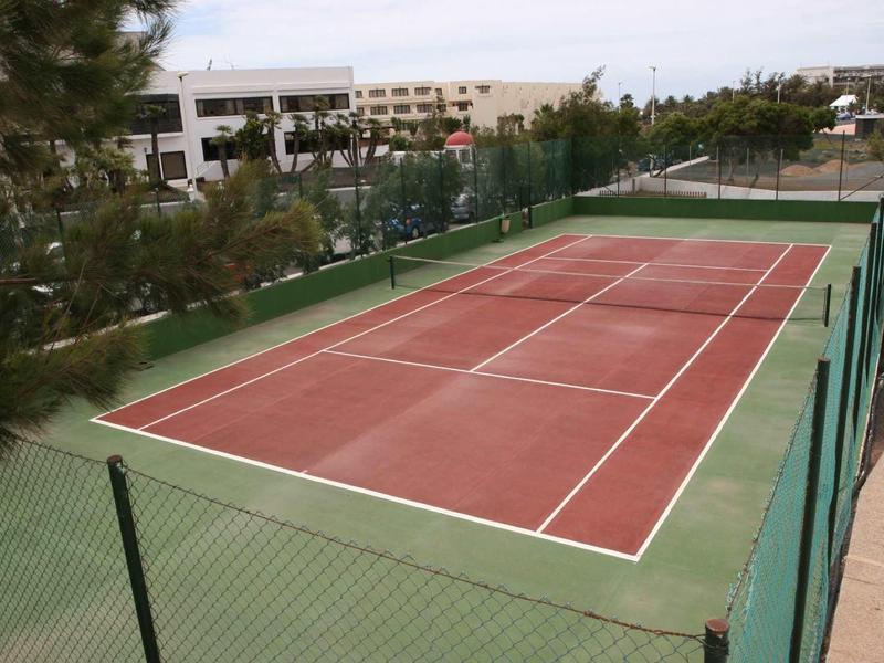 Leerer Tennisplatz mit rotem Spielfeld und grünem Rahmen, umgeben von grünem Maschendrahtzaun.