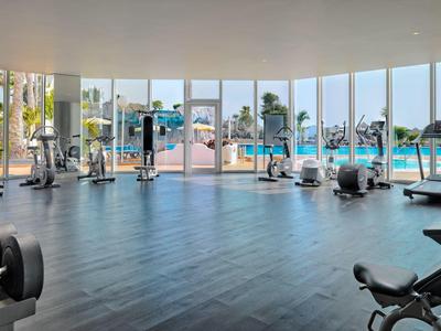 Helles Fitnessstudio mit großen Fenstern, Fitnessgeräten und Blick auf den Pool und Palmen.