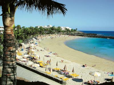 Strand mit gelben Sonnenschirmen, Menschen, Palmen und blauem Meer unter klarem Himmel.