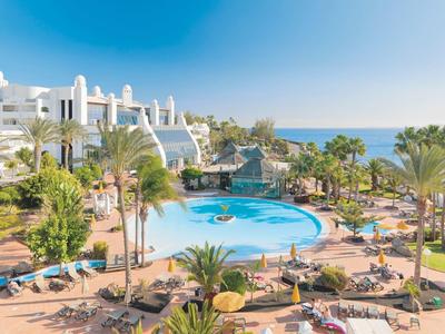 Großer Hotelpool mit Palmen, Liegestühlen und weißem Hotelgebäude unter klarem Himmel.