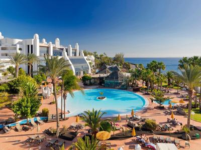 Hotelanlage mit rundem Pool, Palmen und Liegen, im Hintergrund das blaue Meer und klarer Himmel.