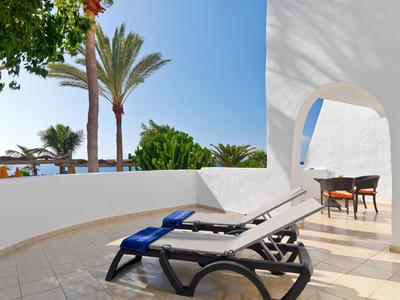 Terrasse mit zwei Liegen, weißer Wand, Palmen und blauem Himmel im Hintergrund.