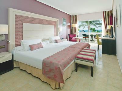 Hell eingerichtetes Hotelzimmer mit zwei Betten, Sitzbank, Schreibtisch und großem Fenster mit Außenblick.