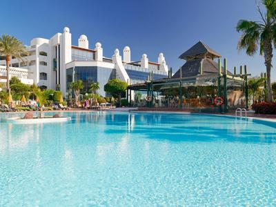 Großer Hotelpool mit klarem blauem Wasser, umgeben von Palmen und weißer Hotelanlage unter blauem Himmel.