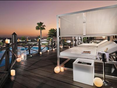 Freiluft-Lounge mit weißen Betten, Holzsteg, Palmen und beleuchtetem Pool bei Sonnenuntergang.