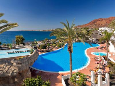 Großer blauer Pool neben weißen Gebäuden, Palmen und Berg, mit Meer im Hintergrund unter klarem Himmel.
