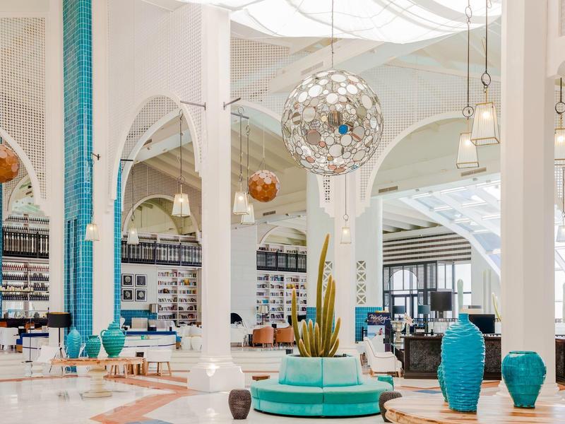 Amplio vestíbulo de hotel con columnas blancas, decoración en tonos azules y lámparas colgantes.