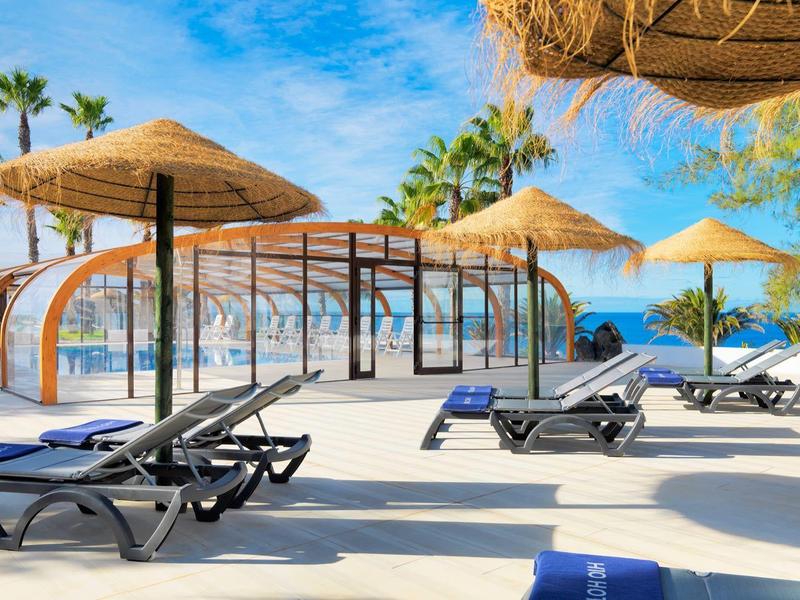 Terraza soleada con tumbonas y sombrillas de paja, palmeras y vista al mar en el hotel.