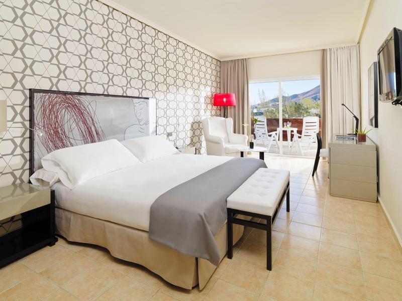 Habitación de hotel luminosa con cama doble, balcón y vista a las montañas durante el día.