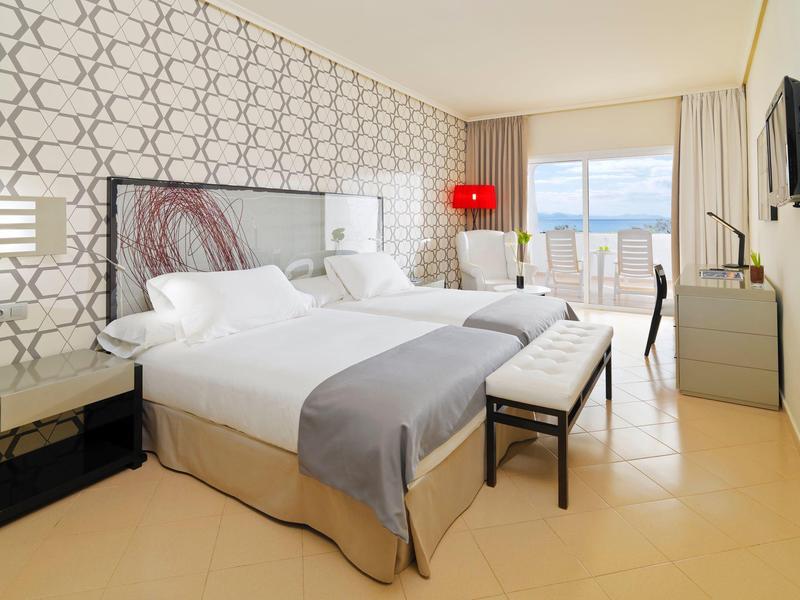 Habitación de hotel luminosa con dos camas, decoración moderna y vista al mar a través de grandes ventanas.