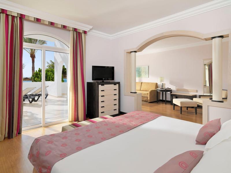 Habitación de hotel luminosa con cama grande, balcón con vista al jardín y área de estar adjunta.