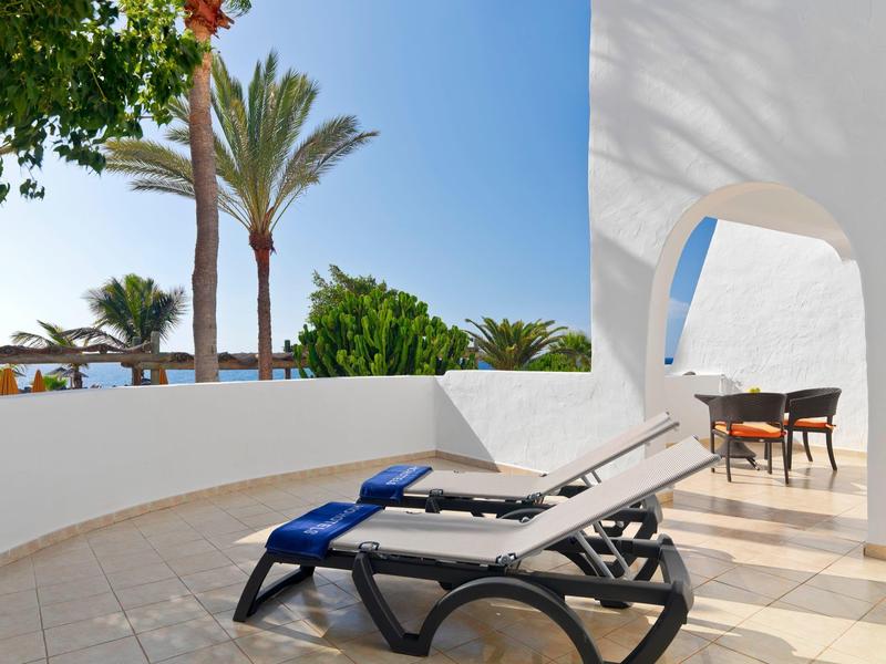 Dos tumbonas con toallas azules en una terraza con vista a palmeras y mar.