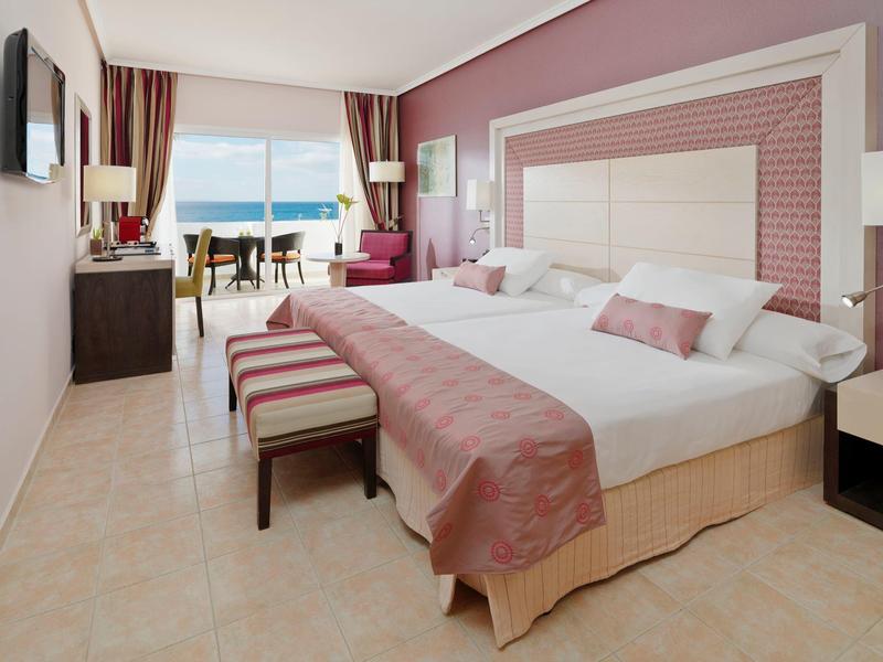 Habitación de hotel elegante con dos camas, acentos rosas y vista al mar y balcón.