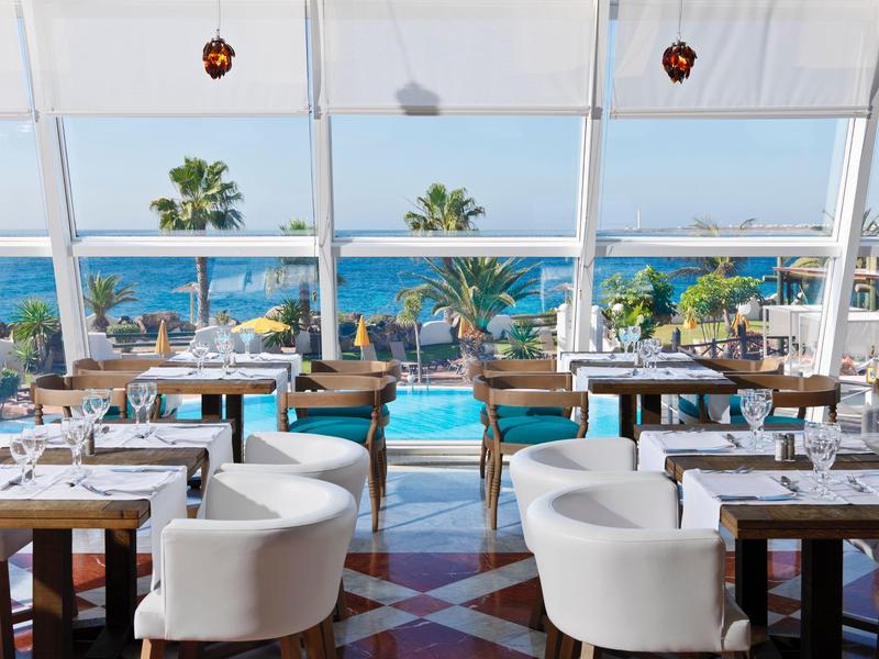 Restaurante iluminado con vista al mar, sillas blancas y mesas de madera junto a la piscina.