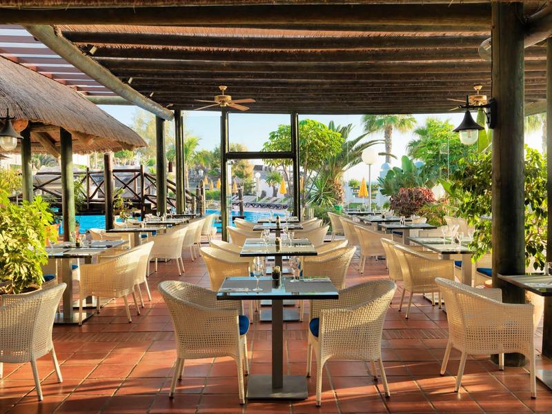 Restaurante al aire libre con sillas y mesas blancas bajo techo de madera, rodeado de plantas verdes con vista al agua.