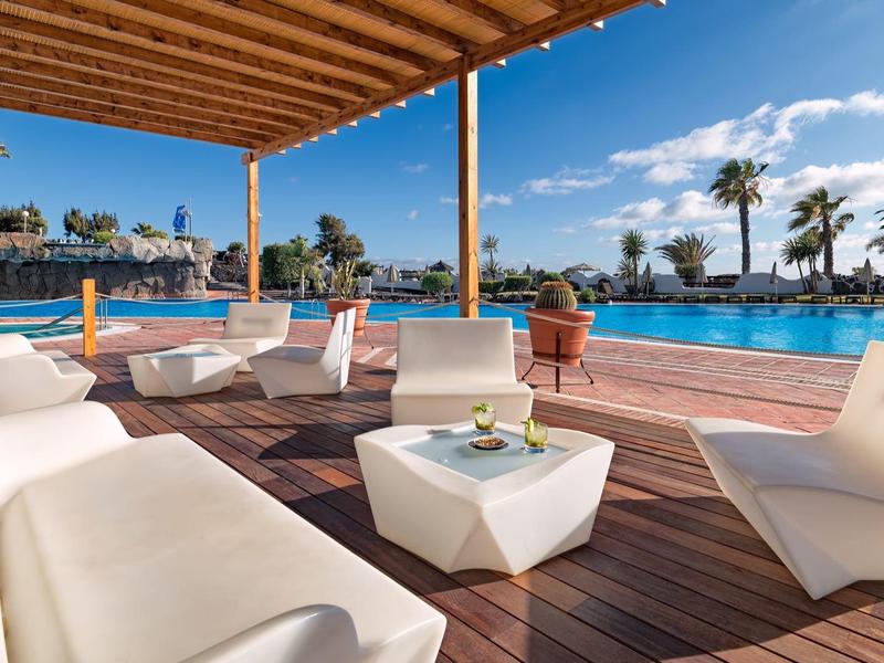 Área de estar con muebles blancos modernos en terraza de madera junto a piscina del hotel bajo cielo azul.