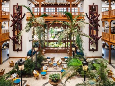 Ampia hall dell'hotel con palme, mobili e balcone aperto al piano superiore.