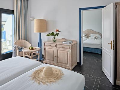 Camera d'hotel luminosa con letto matrimoniale, sedia, comò, balcone e camera adiacente.