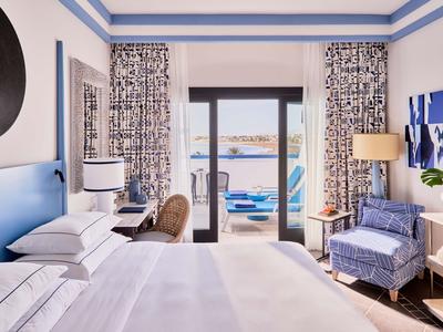 Chambre d'hôtel lumineuse avec lit double, bureau, fauteuil et vue sur terrasse et mer.