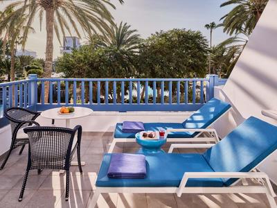 Terrasse avec chaises longues bleues et une table avec des collations, entourée de palmiers.