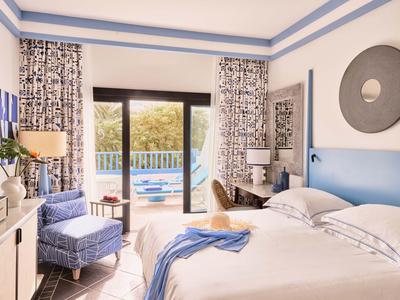 Chambre d'hôtel confortable avec lit double, fauteuil bleu et vue sur la piscine par une porte vitrée.