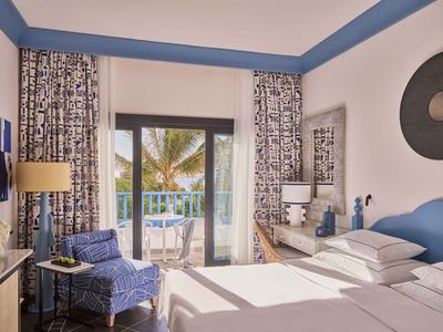 Chambre d'hôtel lumineuse avec deux lits, fauteuil bleu, balcon et vue sur des palmiers.