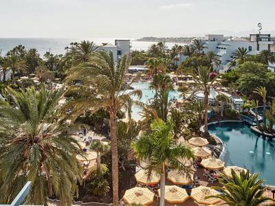 Vue sur une piscine d'hôtel tropicale avec des palmiers, des parasols et une architecture blanche.