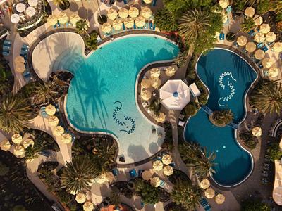 Luftaufnahme eines Hotelpools mit Sonnenschirmen und Palmen in tropischer Umgebung.