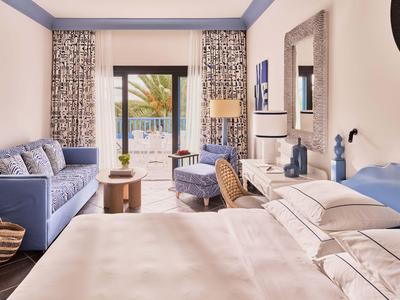 Helles Hotelzimmer mit Doppelbett, Sofa, Sessel und Blick auf eine Terrasse mit Pool.