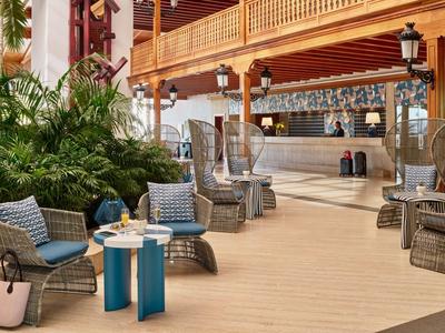 Innenaufnahme einer Hotel-Lounge mit Korbsesseln, kleinen Tischen und Pflanzen in warmem Licht.