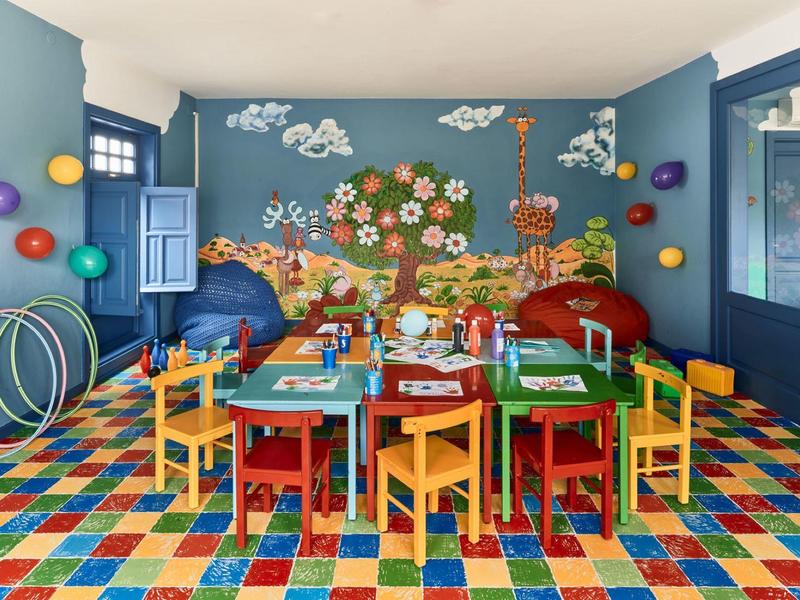 Stanza gioco colorata con piccoli tavoli, sedie e decorazioni murali di animali e fiori.