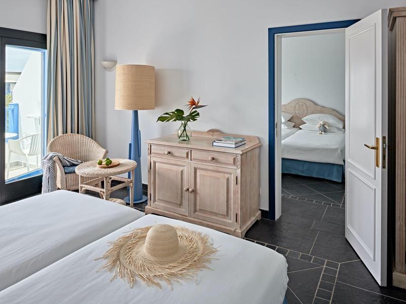 Camera d'hotel luminosa con letto matrimoniale, sedia, comò, balcone e camera adiacente.