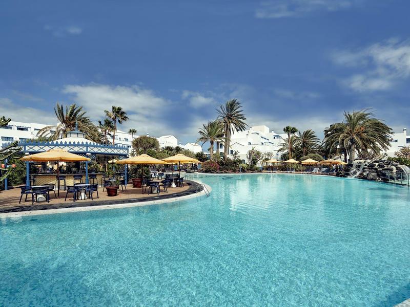 Grande piscina dell'hotel con palme, ombrelloni e aree relax sotto un cielo azzurro