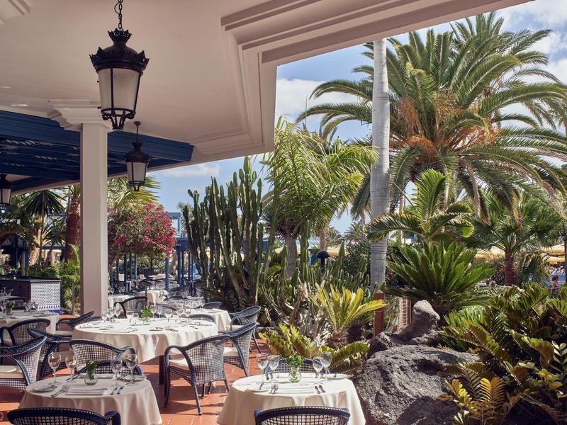 Ristorante all'aperto con tavoli apparecchiati e giardino tropicale con palme e cactus.
