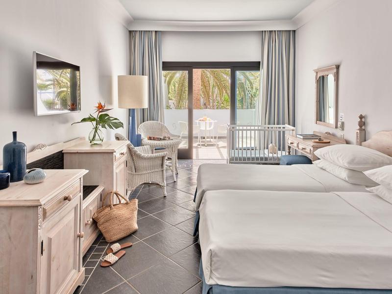 Camera d'hotel luminosa con due letti, scrivania, TV e balcone con vista sulle palme.