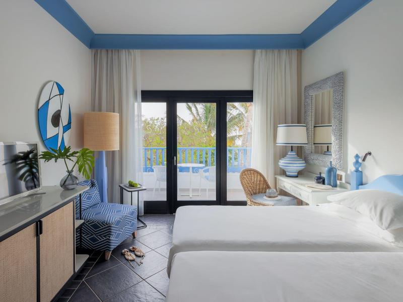 Camera d'hotel luminosa con due letti, accesso al balcone e arredi moderni in tonalità di blu e beige.