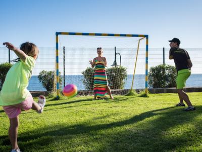 Three children playing soccer on green grass by the sea in sunlight.