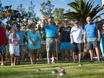 Group of older people playing pétanque outdoors in a sunny park with palm trees.