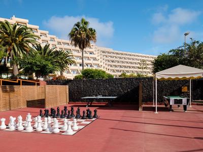 Large outdoor chess set on red ground with palm trees and hotel in the background.