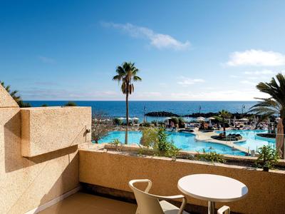 Balkon mit Tisch und Stühlen, Blick auf Pool, Palmen und Meer unter blauem Himmel.