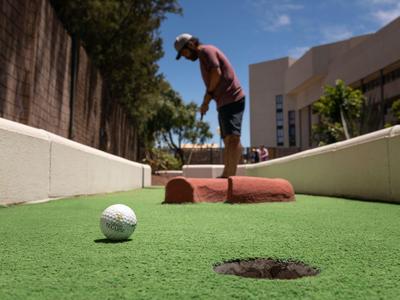 Man playing mini golf on a sunny course with hotel building in the background.