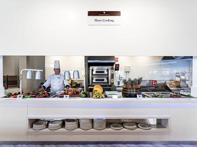 Küche mit Koch, weißem Tresen, großen Töpfen, Pfannen und frischem Gemüse auf der Arbeitsfläche.