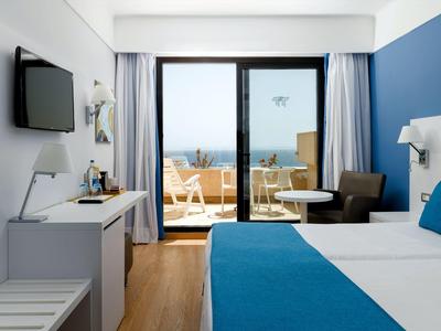 Modernes Hotelzimmer mit blau-weißem Bett, Schreibtisch und Balkon mit Meerblick.