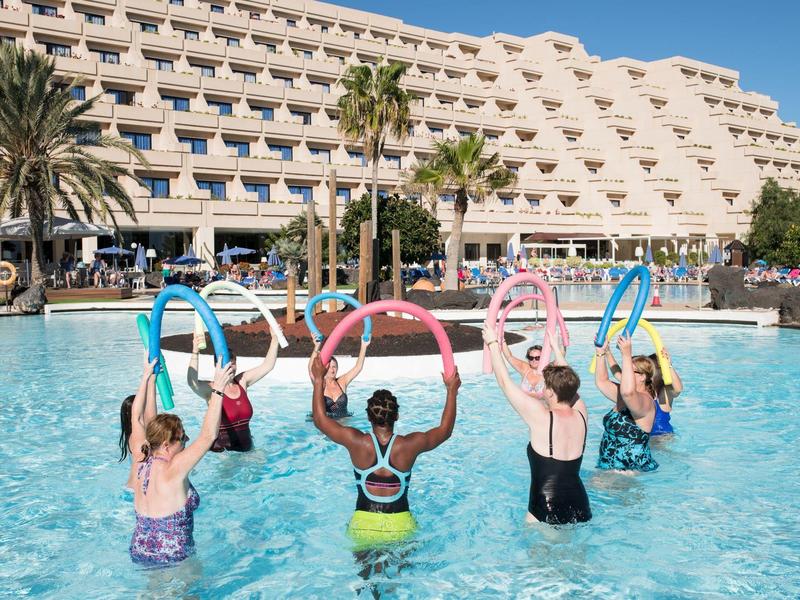 Menschen machen Aquafitness mit bunten Schwimmringen im Pool vor einem großen Hotel in sonniger Umgebung.